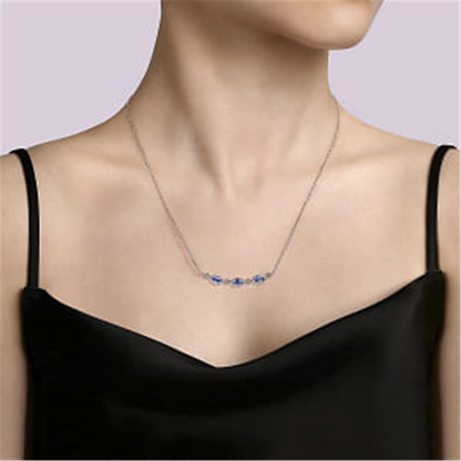 A person wearing a 925 Sterling Silver Bujukan Blue Topaz Bar Necklace from Gabriel & Co, Serial No S174, sits against a light purple background. Adorned with Swiss Blue Topaz gemstones, the necklace beautifully complements their black, sleeveless V-neck top. The focus is on the exquisite necklace and the elegant upper part of the outfit.