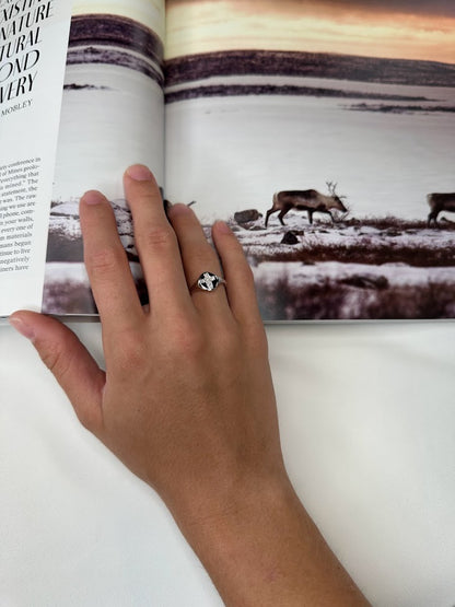 A hand adorned with an Allison Kaufman Diamonds 14K Gold Cross Ring rests on an open book featuring a wildlife photograph with reindeer in a snowy landscape.