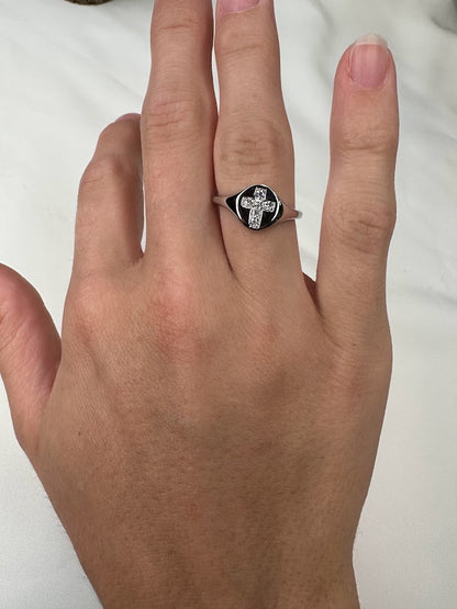 An Allison Kaufman Diamonds 14K gold cross ring, distinguished by its sleek black oval face and silver cross design, rests gracefully on a white surface.