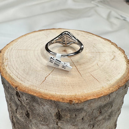 A 14k gold cross ring by Allison Kaufman, featuring a unique design, rests on a round wooden log slice. A size label is attached to the ring, and the wooden surface highlights visible rings and texture. In the background, part of a white fabric can be seen.