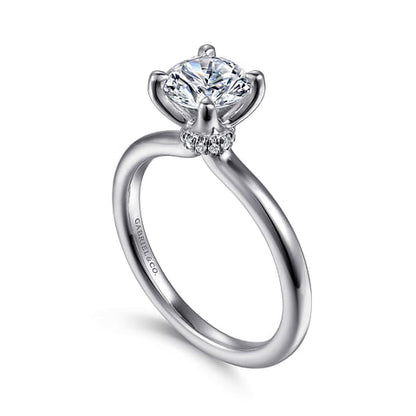 The 14K Gold Diamond Engagement Ring by Gabriel & Co features a customizable, silver engagement ring with a round, brilliant-cut diamond in a prong setting, surrounded by smaller accent diamonds at the base of the main stone. The simple and smooth band enhances the elegance of this stunning 14K white gold ring.