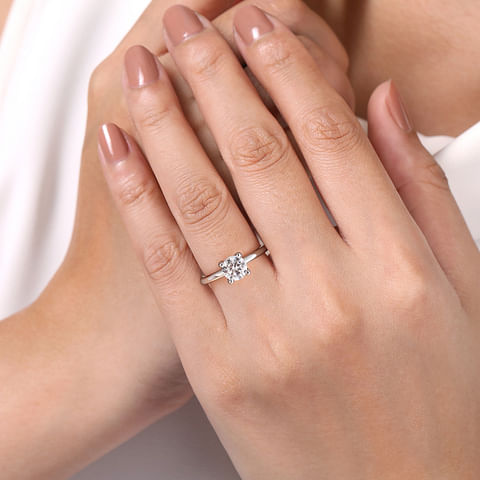 Close-up of a hand with nude-colored nail polish wearing the Gabriel & Co 14K Gold Diamond Engagement Ring featuring a large, square-cut diamond. The hand is held gently by another, with a white fabric in the background.