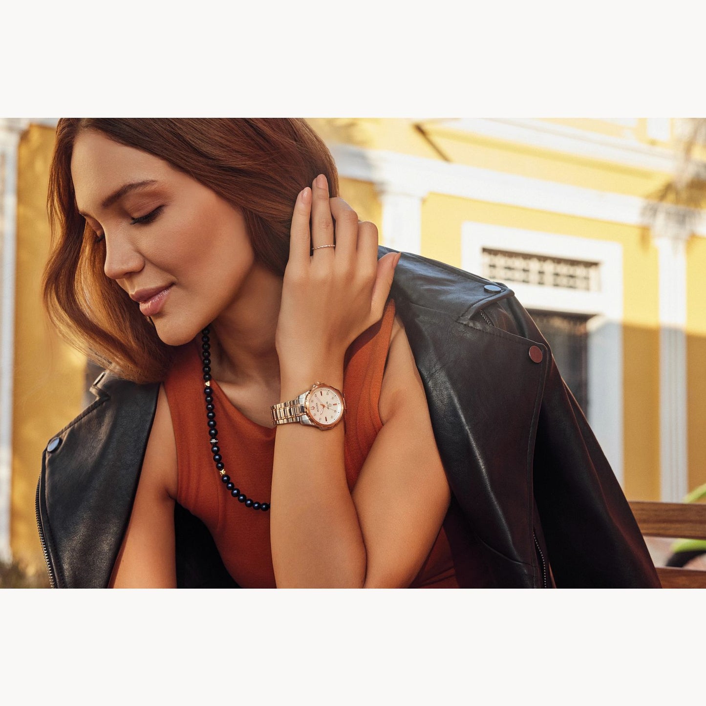 A woman with long brown hair, wearing a black leather jacket draped over her shoulders, an orange top, and a beaded necklace, looks down while resting her chin on her hand. She wears a Bulova Watch Company's Marine Star Ladies' Watch with a Mother of Pearl Dial and diamond accents and is sitting in front of a yellow building.