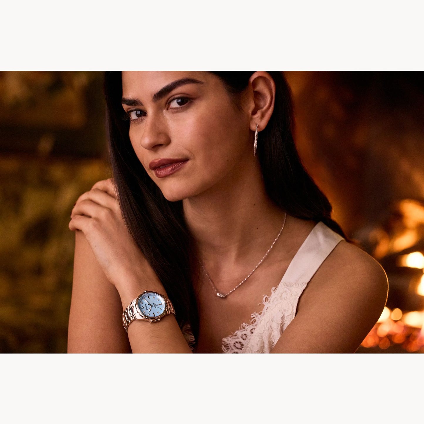 A person with long dark hair is sitting with one hand resting on their opposite shoulder, showcasing a Bulova Marine Star Ladies' Watch with Mother of Pearl and Diamonds. They are wearing a white sleeveless top, drop earrings, and a delicate necklace. The background is softly blurred with warm lighting.