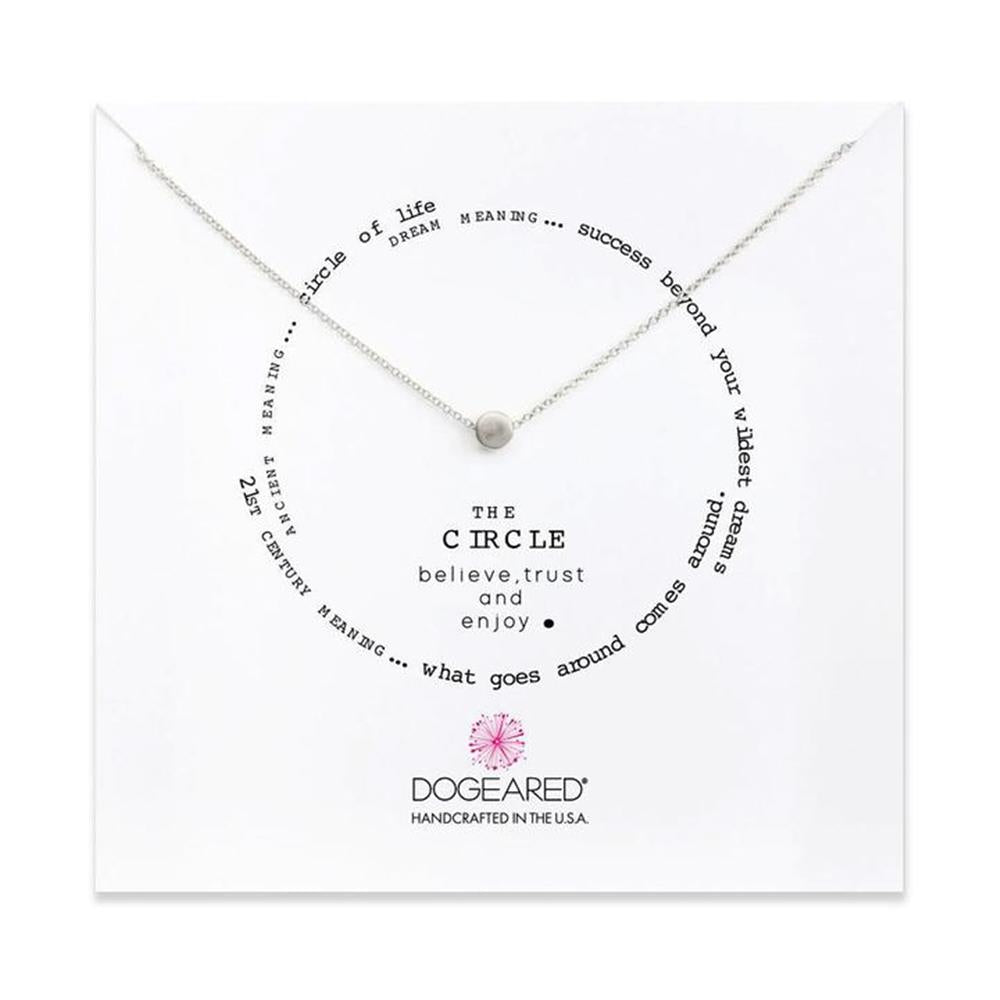 A silver chain necklace with a small circular pendant is displayed on a white card. The card features inspirational quotes arranged in a circle around the necklace, with the text "The Circle Believe, Trust, & Enjoy" in the center. The brand “Dogeared” is at the bottom.