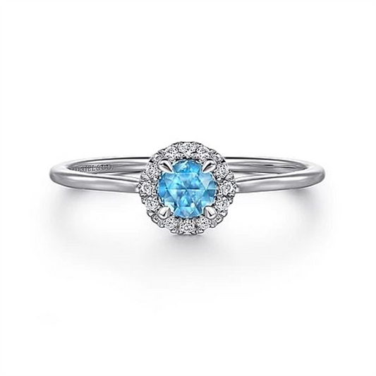 The Gabriel & Co 14K White Gold Blue Topaz and Diamond Halo Promise Ring features a delicate band that emphasizes a dazzling blue topaz at its center, surrounded by a halo of smaller clear diamonds. Displayed against a plain white background, this exquisite ring becomes the highlight of any jewelry collection.