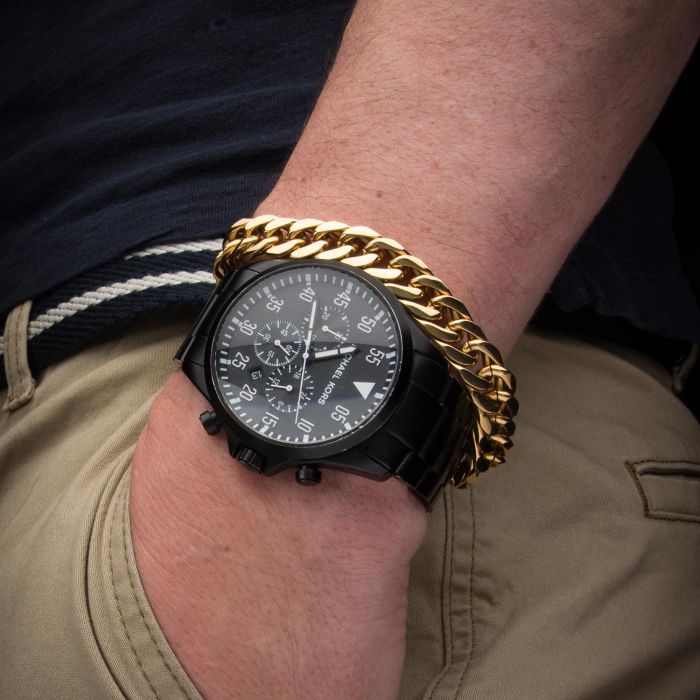 A person wearing a black watch with multiple dials and an INOX Gold Plated Fancy Curb Chain Bracelet on their wrist. The person is also wearing a black shirt and beige pants with a black and white striped belt, and their hand is tucked into their pocket.