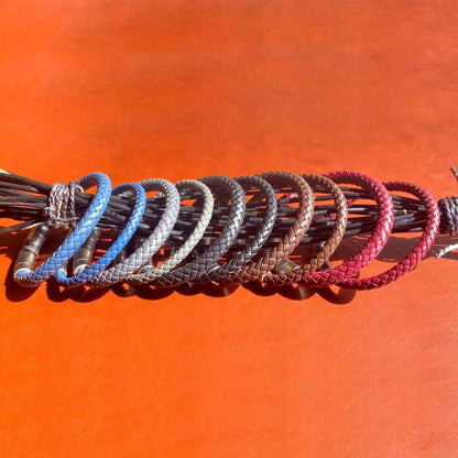 A collection of braided leather bracelets in various colors, including the striking 6mm Blue Genuine Leather Bracelet by INOX, as well as brown, black, red, and other shades in between. Displayed on a wooden branch against a vibrant orange background, the bracelets are neatly arranged from left to right.