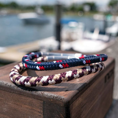 Two intricately woven nylon cord bracelets from INOX lie on a wooden box, with a blurred marina in the background. One bracelet features red, white, and blue colors and has a brushed tubular clasp, while the other is a 6mm Brown, Beige and Dark Brown Nylon Cord Bracelet with an 8.5-8" length.