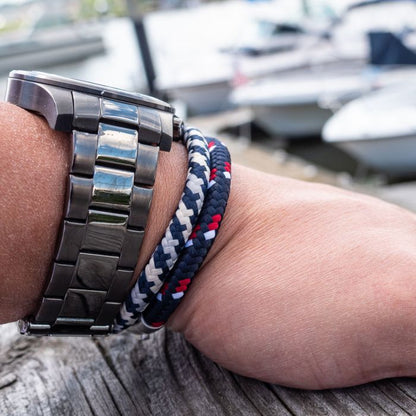 Close-up of a wrist adorned with a stainless steel watch and INOX's 6mm Blue, White, and Red Nylon Cord Bracelet, measuring 8-8.5 inches. The background shows a blurred view of moored boats at a marina on a sunny day.