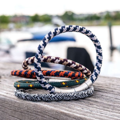 Five braided rope bracelets with metal clasps are displayed on a wooden surface, featuring the 6mm Green, Brown and Yellow Nylon Cord Bracelet by INOX. These vibrant bracelets come in various patterns and colors including blue, orange, and green. The background features an out-of-focus outdoor scene with greenery and water.