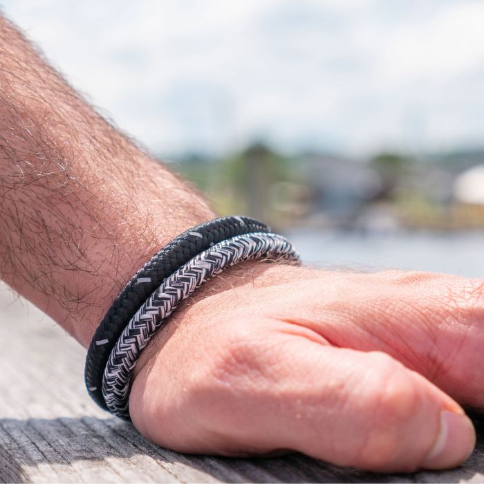 Close-up of a person’s hand resting on a wooden surface, adorned with two men's INOX stainless steel bracelets—one black and the other featuring the 6mm Black and White Nylon Cord Bracelet (8.5-8")—against a blurred background of a waterfront setting with boats and a cloudy sky.