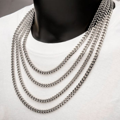 A person wearing a white t-shirt displays several layers of thick, silver men's necklaces around their neck. The 6mm Steel Miami Cuban Chains by INOX, crafted from stainless steel, feature a polished, interlocking link design and drape elegantly over the shirt, creating a multi-layered, stylish appearance.