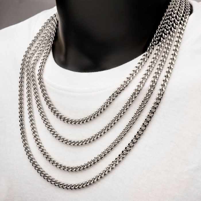 Close-up of a person's neck wearing multiple layered men's necklaces over a white T-shirt. A standout 6mm Steel Miami Cuban Chain by INOX, measuring 22 inches, along with other stainless steel chains of varying lengths are neatly draped, creating a bold and stylish look. The neutral background highlights the jewelry and garment perfectly.