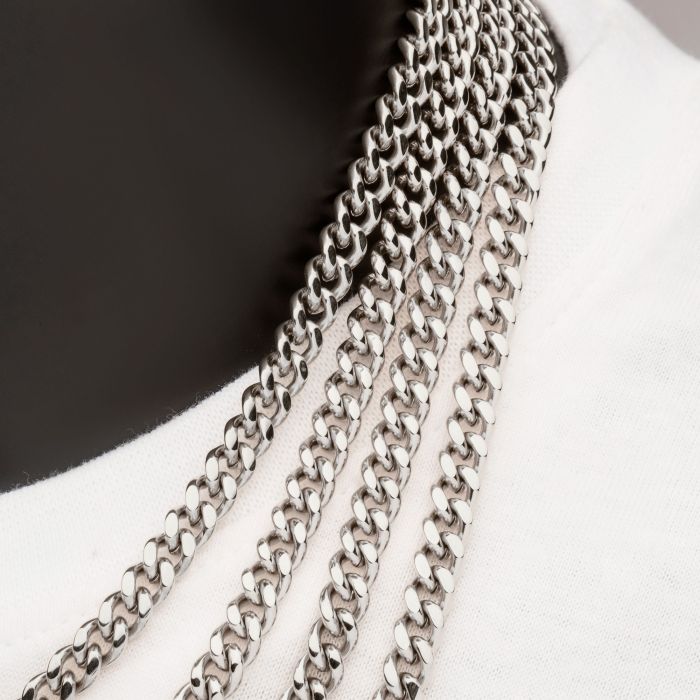 Close-up of a person wearing multiple strands of silver chain necklaces, including the 6mm Steel Miami Cuban Chain by INOX and stainless steel chains, over a white garment. The chains are evenly spaced and feature classic link designs, creating a layered and stylish look. The background shows part of the person's neck and shoulder.