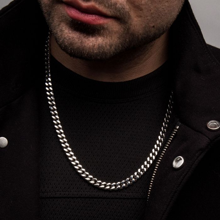 Close-up of a man wearing the 6mm Steel Miami Cuban Chain necklace by INOX. He is dressed in a black jacket with silver snaps and a black textured shirt underneath. The background is plain grey. His stubble-covered chin and part of his mouth are visible.