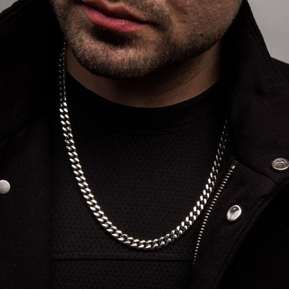 Close-up of a person wearing a dark jacket and shirt, showcasing the 6mm Steel Miami Cuban Chain by INOX around their neck. The person has a short beard, and their lips are slightly parted. The background is neutral.