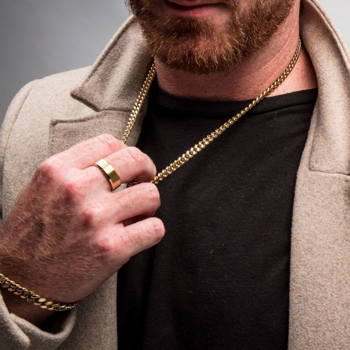 A close-up of a person with a red beard wearing a beige coat over a black shirt. They have an INOX 6mm 18K Gold Plated Miami Cuban Chain | 22" around their neck, which they are holding with their hand. The same hand is adorned with a gold bracelet and a large gold ring.