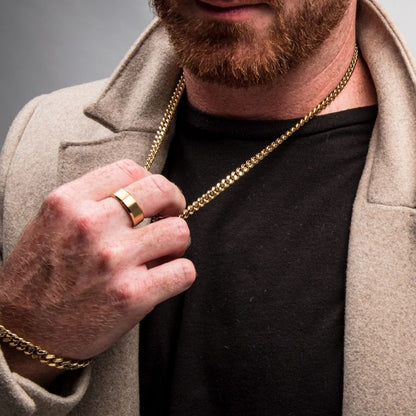 Close-up of a man wearing a beige coat over a black shirt. He has a trimmed beard and is holding an 8mm 18K Gold Plated Miami Cuban Chain by INOX. He is also wearing a gold bracelet and a gold ring on his finger.