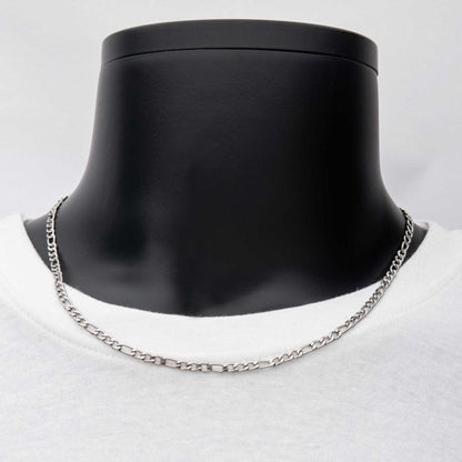 A close-up of the 4mm Steel Figaro Chain from INOX, elegantly displayed on a black mannequin bust. The men's necklace rests on a white shirt, creating a striking contrast between the metallic shine of the 20-inch stainless steel chain and the black and white background. The focus is entirely on this exquisite piece by INOX.