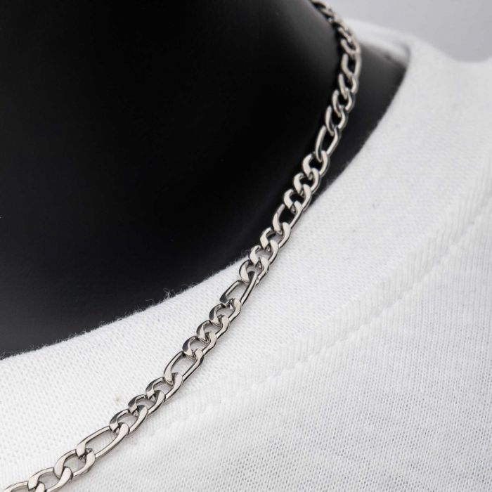 A close-up image of the 4mm Steel Figaro Chain | 20" by INOX worn around the neck, displayed over a plain white shirt. The men's necklace highlights an intricate pattern of interconnected links, emphasizing the texture and detail of the stainless steel jewelry. The background is a neutral shade.