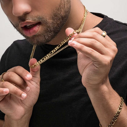 A person with facial hair, wearing a black shirt, holds up an INOX 6mm 18K Gold Plated Figaro Chain necklace that measures 24 inches. They are also adorned with several gold rings and another gold chain around their neck. Their lips are slightly parted as they gaze at the chain in their hand.