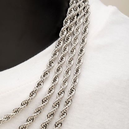 Close-up of several INOX 6mm Steel Rope Chains (22") neatly arranged in parallel, draped over a white fabric. The chains exhibit intricate twisting patterns and reflect light, highlighting their metallic shine. The white fabric serves as a simple backdrop.