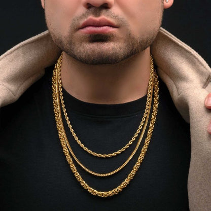 A close-up of a man wearing three INOX 6mm 18K Gold Plated Rope Chains of varying lengths over a black shirt. His hands are adjusting a beige jacket draped over his shoulders. Only the lower half of his face, including a trimmed beard, is visible. The background is solid black.