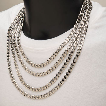 A close-up image of a person wearing a white shirt and multiple layered INOX 8mm Steel Bevel Curb Chains. The stainless steel chains, including the 20" INOX chain, are of varying lengths, creating a cascading effect across the person's chest. The plain background highlights the men's necklaces.