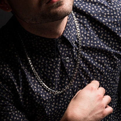 Close-up of a person wearing a blue patterned button-up shirt and the INOX 8mm Steel Bevel Curb Chain necklace. The individual's left hand is placed over their chest, partially covering a button on the shirt. The dark background emphasizes both the shirt and this bold men's necklace.