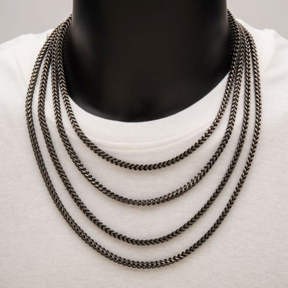 Close-up of a black mannequin wearing a white shirt and displaying an INOX 4mm Oxidized Steel Franco Chain, measuring 20 inches in length. The dark, metallic links are arranged in layered strands that lie flat against the chest, creating a striking effect from shorter to longer lengths.