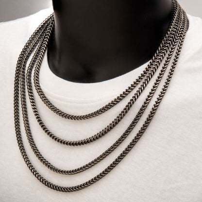 A person is wearing a white shirt with multiple layered 4mm Oxidized Steel Franco Chains from INOX draped around their neck. The chains, which are 20 inches long, vary in length and create an overlapping effect. The background features a close-up of the chest and neck area only.