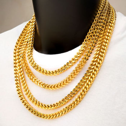 Close-up of a person wearing multiple 4mm 18K Gold Plated Franco Chains by INOX, each of varying lengths, layered over a white shirt. The thick gold-plated chains have a shiny, reflective surface. The neutral background does not distract from the jewelry.