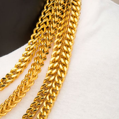 Close-up of three INOX 4mm 18K Gold Plated Franco Chains, each 20 inches long, resting on a white fabric. The chains are shiny and reflective, showcasing a detailed and elegant pattern. The background is partially black, providing a striking contrast to highlight the gold chains.