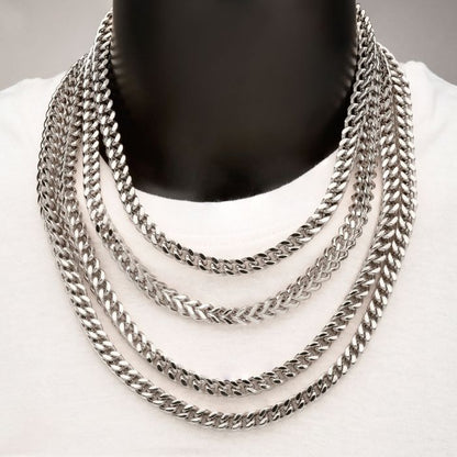 A person wearing a white shirt with four INOX 6mm Steel Franco Chains of varying lengths layered around their neck. The 20" necklaces have a robust, intricate design, and lay flat against the chest, creating a striking, layered look against the plain background.