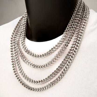 A person wearing a white shirt is adorned with multiple thick, silver chain necklaces of varying lengths, including the sleek 6mm Steel Franco Chain by INOX. The chains are layered, creating a stylish and bold fashion statement. The neutral background emphasizes the shiny texture of the men's stainless steel necklace collection from INOX.