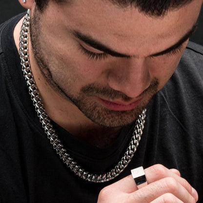 A person with a closely trimmed beard and dark hair looks down while wearing an INOX 6mm Steel Franco Chain necklace (24") and holding a square-shaped silver ring close to their face. The background is dark, and the individual is dressed in a black shirt.