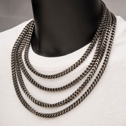 A person wearing a white shirt adorned with four layered silver chain necklaces, including the 6mm Oxidized Steel Franco Chain by INOX. The chains, crafted from stainless steel, vary in length to create a cascading effect around the neck. The plain and neutral background enhances the appeal of these men's necklaces.