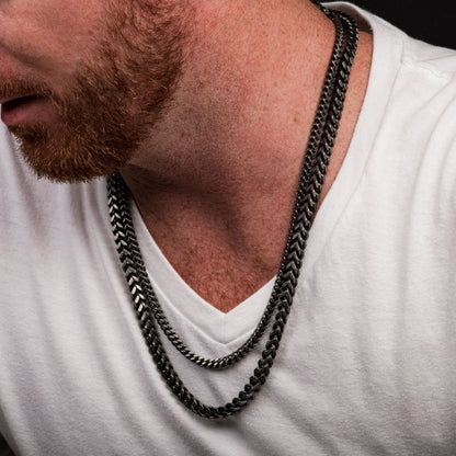 A person with a reddish beard is wearing a white V-neck shirt and two black metal chain necklaces. One, a 6mm Oxidized Steel Franco Chain by INOX, and the other a stainless steel necklace, are of different lengths and both rest over the shirt. The person's face is partially out of the frame.