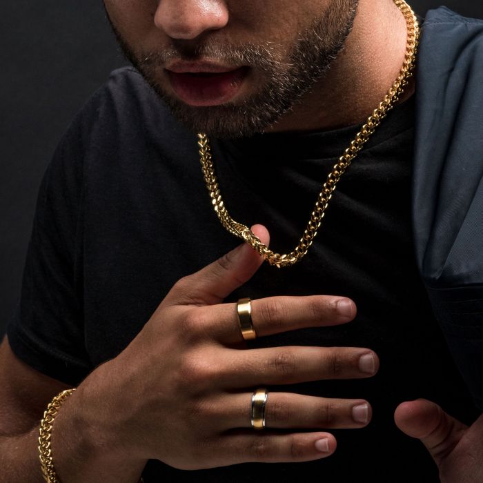 Close-up of a person wearing a black shirt and accessorized with 18K gold plated jewelry, including two thick INOX men's necklaces—a 6mm 18K Gold Plated Franco Chain at 24 inches—around their neck, two gold rings on their right hand, and a stainless steel bracelet on their left wrist. The person is touching the chains with their right hand.