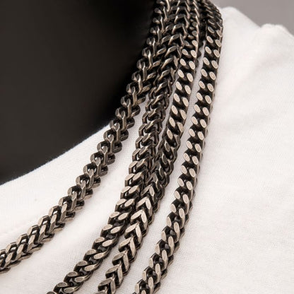 Close-up of multiple layered 8mm Oxidized Steel Franco Chains by INOX and stainless steel men's necklaces worn around the neck against a white shirt background. The chains feature a textured and reflective finish, creating an eye-catching display.