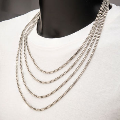 Close-up of a person wearing a white shirt, showcasing multiple layers of silver chains around their neck. Among them, an 8mm Diamond Cut Curb Chain Necklace from INOX stands out—a 24" chain that glistens against the white fabric. The image highlights its intricate design and shine while the person's face remains unseen.