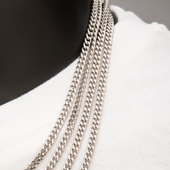 A close-up image of a person wearing a white shirt and multiple silver chain necklaces, including the 8mm Diamond Cut Curb Chain Necklace, 24" from INOX, draped around their neck. The neutral background emphasizes the shiny texture and intricate links of the Men's Stainless Steel Necklace collection by INOX.