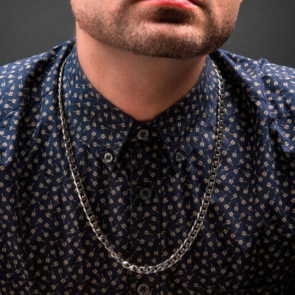 Close-up of a person wearing a navy blue button-up shirt with an abstract pattern, and INOX's 8mm Diamond Cut Curb Chain Necklace. The 24" stainless steel necklace gleams silver around their neck. The person's face is partially out of frame, showing only the chin and part of the beard against a solid black background.