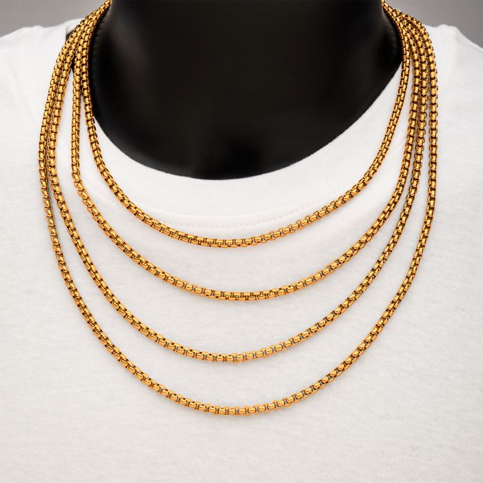 A person wearing a white t-shirt and multiple layers of 18K gold plated chain necklaces, including the INOX 4mm 18K Gold Plated Bold Box Chain Necklace, which measures 20 inches. The necklaces cascade elegantly around the neck. The neutral background highlights the men's stainless steel necklace and other jewelry pieces beautifully.