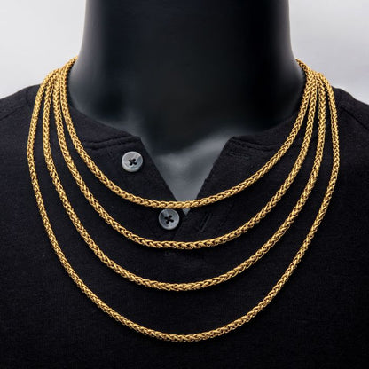 A person wearing a black buttoned shirt with five INOX 4mm 18K Gold Plated Wheat Chain Necklaces, each 20 inches long and of varying lengths around their neck. The men's necklaces are layered and evenly spaced, displaying a stylish and elegant look. The background is plain and neutral.