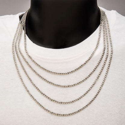 A person wearing a white shirt is adorned with the 4mm Steel Classic Curb Chain from INOX, a men's stainless steel necklace. The 20" silver chains create a cascading effect as they drape over the chest. The plain background draws full attention to the neck and upper chest area.