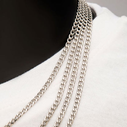 A close-up shot of a person wearing multiple INOX 6mm Steel Classic Curb Chains around their neck. The 22" men's necklaces are finely woven and rest on a white fabric, likely a shirt. The dark background creates a striking contrast with the silver of the chains and the white fabric.