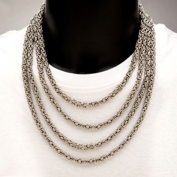 A close-up image shows a person wearing a white shirt and the 6mm Steel King Byzantine Chain by INOX. The intricate design of the stainless steel chains drapes elegantly across the chest, creating a statement piece that stands out against the white fabric.