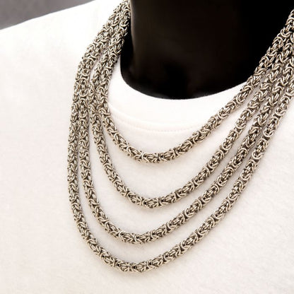 A close-up of a mannequin wearing the INOX 6mm Steel King Byzantine Chain, layered elegantly over a plain white shirt. The 22" stainless steel chain features an intricately braided pattern that stands out strikingly against the simplicity of the shirt.